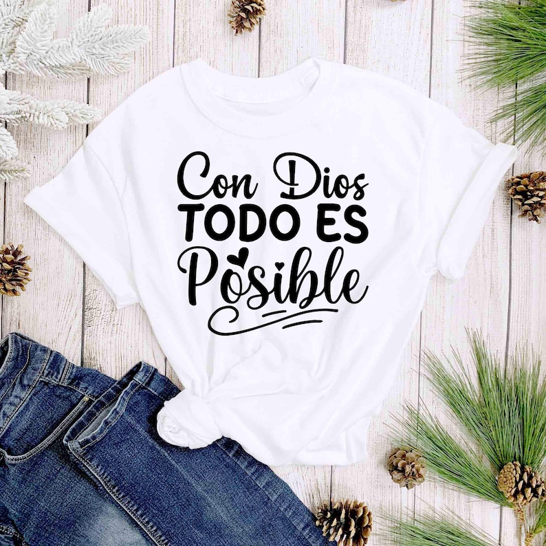 Puede incluir: Camiseta blanca con la frase "Con Dios TODO ES Posible" en escritura negra. La camiseta se combina con vaqueros azules y est&aacute; decorada con pi&ntilde;as y vegetaci&oacute;n.