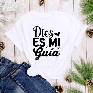 Puede incluir: Una camiseta blanca con la frase "Dios Es Mi Gu&iacute;a" en escritura negra. La camiseta se combina con vaqueros azules. La imagen est&aacute; decorada con agujas de pino y pi&ntilde;as, lo que sugiere un tema de temporada.