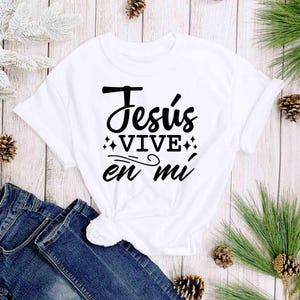 Puede incluir: Camiseta blanca con el texto "Jes&uacute;s vive en m&iacute;" en escritura negra. La camiseta se combina con vaqueros azules. La imagen est&aacute; decorada con agujas de pino y pi&ntilde;as sobre un fondo de madera clara.