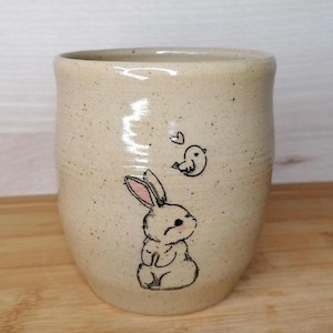 Könnte beinhalten: Eine beige Keramiktasse mit gesprenkeltem Glase zeigt eine handgezeichnete Illustration eines weißen Kaninchens mit rosa Ohren und eines kleinen Vogels mit einem rosa Herzen darüber.