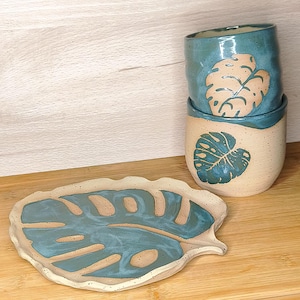 Puede incluir: Un juego de cerámica hecho a mano con una combinación de colores turquesa y beige. El juego incluye dos tazas apiladas con diseños de hojas y un plato a juego con forma de hoja. Las tazas y el plato están sobre una superficie de madera.