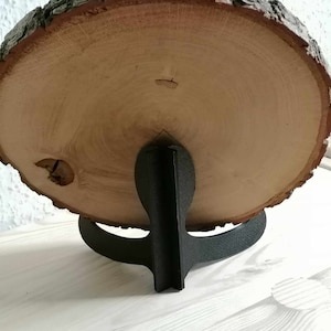 Könnte beinhalten: Eine runde Holzscheibe mit natürlicher Rindenkante, montiert auf einem schwarzen Metallständer. Das Holz ist hellbraun und hat eine glatte Oberfläche.