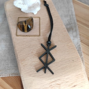 Könnte beinhalten: Ein schwarzer Anhänger mit einem geometrischen Bindrunen-Symbol an einer schwarzen Kordel. Der Anhänger wird zusammen mit einer kleinen weißen Figur einer sitzenden Person und einem polierten braun-goldenen Tigerauge auf einem hellen Holzblock präsentiert.