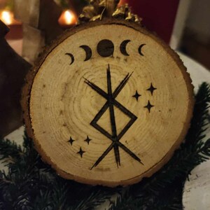 Könnte beinhalten: Ein runder Holzschmuck mit einem schwarz-weißen Symbol und drei Mondsicheln. Das Symbol ist ein doppelseitiges, diagonales Kreuz mit einem Quadrat in der Mitte. Über dem Symbol befinden sich drei kleine Sterne.