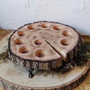 Könnte beinhalten: Ein Holztablett mit einem natürlichen Rindenrand und acht kleinen, runden Vertiefungen. Das Tablett wird von drei dekorativen Metallfüßen getragen.