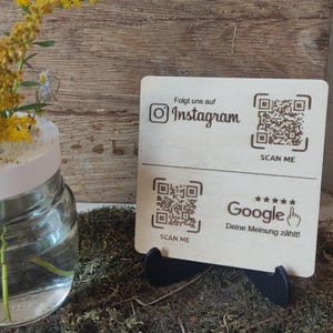 Könnte beinhalten: Ein Holzschild mit zwei QR-Codes. Der obere QR-Code lautet "Scan Me" und "Folgt uns auf Instagram". Der untere QR-Code lautet "Scan Me" und "Google Deine Meinung zählt!"