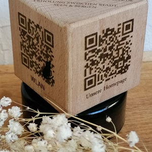 Könnte beinhalten: Ein Holzwürfel mit einem QR-Code und dem Text "SCHNEELE FERIENWOHNUNGEN WWW.FERIENWOHNUNGEN-SCHNEELEDE ERHOLUNG ZWISCHEN STADT SEEN & BERGEN" auf der Oberseite. Der Würfel steht auf einem schwarzen Sockel und im Vordergrund befindet sich eine kleine weiße Blüte.