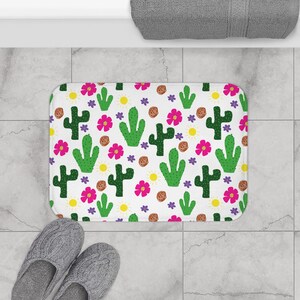 May include: White bath mat with a repeating pattern of green cacti, pink flowers, and yellow suns.