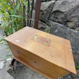 May include: A vintage wooden Kodak Film Tank box with a rectangular shape and a weathered, aged appearance. The box features the Kodak logo in red and green on the top. The box is resting on a weathered wooden surface.