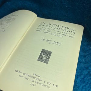 May include: An open hardcover book with cream-colored pages featuring the title An Alphabetical Encyclopedia of Institutions, Persons, Events, Etc., of Ancient History and Geography by Dr. Emil Reich, published in London and New York in 1906.