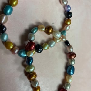 May include: A necklace composed of irregularly shaped, colorful pearls. The pearls are in shades of red, black, gold, blue, and cream. The necklace is displayed on a light pink surface.