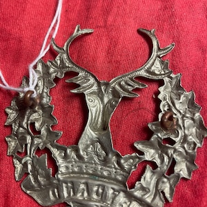 May include: A silver metal badge featuring a deer's head at its centre, encircled by a wreath of leaves. The deer has large antlers and a crown above its head. The words "The Royal" are inscribed on a banner beneath the deer.