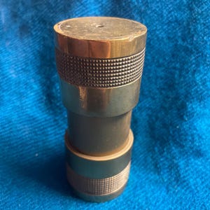 May include: A brass bottle stopper with a textured band. The stopper has a cylindrical shape with a flat top and a smaller base. The brass has a warm, golden hue, and the textured band adds a decorative element.