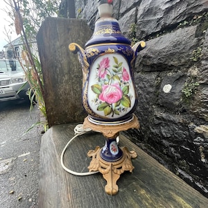 Puede incluir: Lámpara de mesa de porcelana vintage con base azul oscuro y detalles dorados. La lámpara presenta un diseño floral con rosas rosas y hojas verdes. Se apoya sobre una base decorativa de latón y tiene un cable blanco.