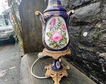 May include: A vintage porcelain table lamp with a dark blue base and gold accents. The lamp features a floral design with pink roses and green leaves. The lamp sits on a decorative brass base and has a white cord.