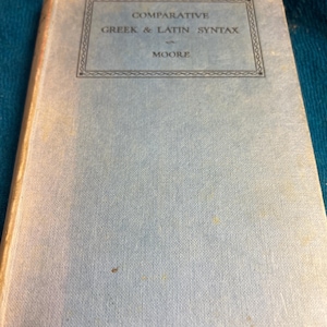 May include: A light blue hardcover book titled Comparative Greek & Latin Syntax by Moore. The front cover features a rectangular border with decorative line work surrounding the text. The book has a textured fabric finish and a light blue spine.