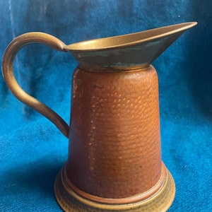 May include: A copper pitcher with a hammered texture, featuring a brass handle and rim. The pitcher has a wide base and a spout for pouring. The overall color is a warm copper with brass accents, creating a vintage aesthetic.