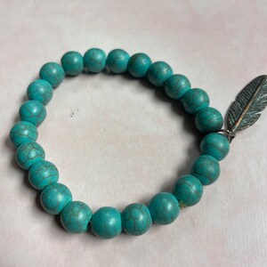 May include: A turquoise beaded bracelet with a silver feather charm. The beads are round and have a textured surface.