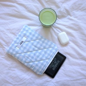 May include: A blue and white gingham quilted tablet sleeve with a zipper closure. The sleeve has a label that says "Daisy" on it. A black e-reader with the text "Sunrise on the Reading" is inside the sleeve. A white wireless charging case and a glass of green liquid are on a white bedspread.