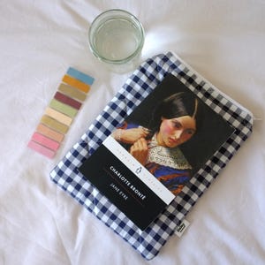 May include: A copy of "Jane Eyre" by Charlotte Brontë, with a portrait of a woman on the cover, rests on a blue and white checkered pouch. A glass of water and a strip of colorful sticky notes are also present.