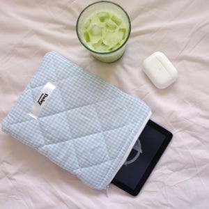 Könnte beinhalten: Eine hellblaue, gesteppte Tablet-Hülle mit Vichy-Karo-Muster und einem weißen Bandetikett mit der Aufschrift "Daisy". Ein Glas grünes Getränk mit Eis und ein weißes kabelloses Kopfhörer-Etui liegen ebenfalls auf einem rosa Laken.