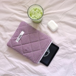 May include: A purple and white gingham tablet sleeve with a white label that says "Daisy". A black tablet is partially visible. A glass of green drink with ice and a white case are also in the image.