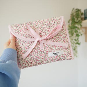 May include: A rectangular fabric pouch featuring a pink and green floral pattern on a white background. The pouch has a light pink ribbon tie closure and a white rectangular fabric label on the front with the black text Daisy Handmade.