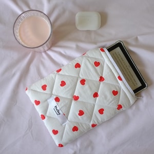 Puede incluir: Una funda blanca acolchada para lector de libros electrónicos con un patrón de corazones rojos, que contiene un lector de libros electrónicos. Un vaso de líquido rosa, una funda blanca y una pequeña etiqueta blanca con la palabra "Daisy" también son visibles sobre una superficie blanca.
