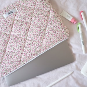 May include: A floral patterned laptop sleeve with a zipper closure. The sleeve is on top of a laptop computer. There are three white pens, a green pen, a tube of pink hand cream, and a bottle of pink lotion on a pink surface.
