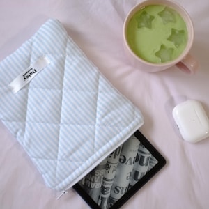 May include: A light blue quilted fabric case with white stripes and a zipper closure. The case has a white label with the text "Daisy" on it. The case is lying on a pink surface with a black e-reader and a white wireless charging case.