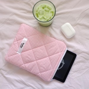 May include: A pink and white gingham quilted tablet case with a white zip, holding a black tablet. A glass of green drink with ice and a white wireless earbud case are also present.