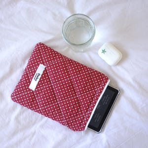 Op de afbeelding: Een rode, gewatteerde tablethoes met witte stippen, met een tablet. Een glas water en een stuk zeep met een ster liggen naast de hoes. De hoes heeft een wit label met de tekst "Baby".