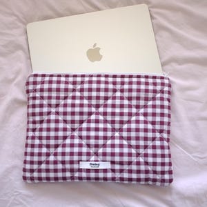 Op de afbeelding: Een gevoerde laptophuls met een gingham-patroon in wit en bordeauxrood. De hoes heeft een wit label met het woord "Daisy" erop gedrukt.