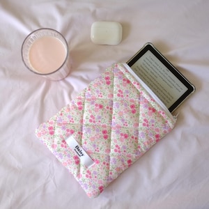 May include: A floral quilted e-reader case with a white zipper, holding an e-reader. A glass of pink liquid and a white bar of soap are also in the image. The case has a small white tag that says "Daisy".