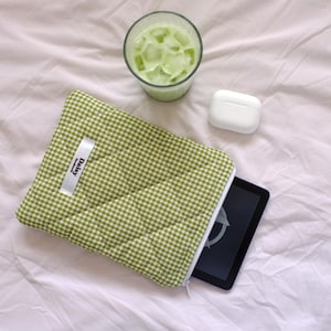 Könnte beinhalten: Eine grün-weiß karierte Tablethülle mit einem weißen Etikett mit der Aufschrift "Daisy". Ein Glas grünes Getränk mit Eis und ein weißes kabelloses Ohrhörer-Etui sind ebenfalls im Bild. Die Artikel liegen auf einem weißen Laken.