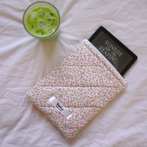 Può includere: Una custodia per e-reader imbottita bianca con stampa floreale rosa e chiusura a zip. La custodia ha un'etichetta bianca con la parola "Daisy" stampata su di essa. La custodia è appoggiata su un copriletto bianco con un bicchiere di liquido verde accanto. L'e-reader è all'interno della custodia e lo schermo mostra il titolo "Sunrise on the Reaping".