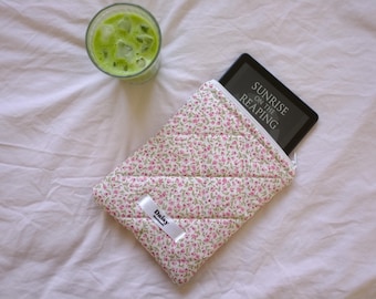 May include: A white quilted e-reader case with pink floral print and a zipper closure. The case has a white label with the word "Daisy" printed on it. The case is lying on a white bedspread with a glass of green liquid next to it. The e-reader is inside the case and the screen shows the title "Sunrise on the Reaping".