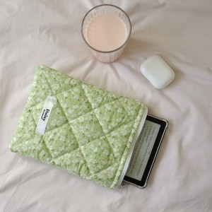 May include: A green floral quilted e-reader case with a white zip, holding an e-reader. A glass of pink liquid and a white bar of soap are next to the case on a white sheet. The case has a white tag that says "Daisy".