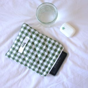 Könnte beinhalten: Eine grün-weiß karierte Handyhülle mit einem weißen Etikett mit der Aufschrift "Daisy" ist geöffnet und zeigt ein Telefon. Ein klares Wasserglas und ein weißes Seifenstück mit einem grünen Stern liegen ebenfalls auf der weißen Oberfläche.