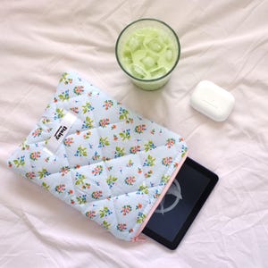 May include: A light blue quilted tablet sleeve with a floral pattern, a pink zip, and a white tag that says "Daisy". A black tablet is partially visible inside the sleeve. A glass of green drink with ice and a white bar of soap are also in the image.