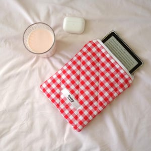 May include: A red and white gingham patterned e-reader case with a white zipper, holding an e-reader. A glass of pink liquid and a white wireless earbud case are also in the image. The case has a white tag that says "Daisy".