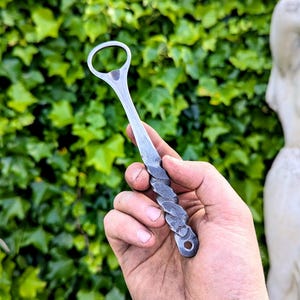 May include: A hand-forged metal bottle opener with a twisted handle design. The opener has a loop at the top for leverage and a small hole at the base of the handle. The background is a green ivy wall.