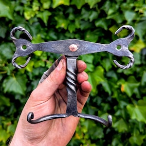 Puede incluir: Gancho de pared de metal forjado a mano con un mango retorcido y extremos rizados decorativos. El gancho es de color gris oscuro con un remache de cobre en el centro. El diseño es rústico e industrial.