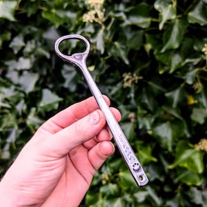 Pode incluir: Um abre-garrafas de metal forjado à mão com um cabo longo e fino, apresentando um acabamento texturizado e um pequeno recorte circular na base. A ferramenta possui uma cabeça arredondada de alça aberta para remover tampas de garrafas.