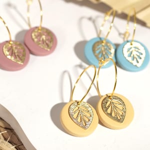 Könnte beinhalten: Drei Paar goldene Creolen mit einem runden, bunten, tonfarbenen Anhänger mit einem goldenen Blattdesign. Die Anhänger sind in rosa, blau und gelb.