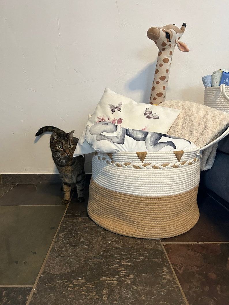 Beiger Aufbewahrungskorb mit Henkeln im Wohnzimmer, gefüllt mit Kissen und Decken. Eine Katze steht links vom Korb.