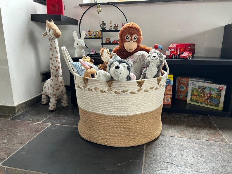 Beiger Aufbewahrungskorb mit Henkeln im Kinderzimmer, gefüllt mit Spielzeug / Plüschtieren. Katze, Hund, Hase, Affe. Im Hintergrund steht noch eine Plüschtier Giraffe.