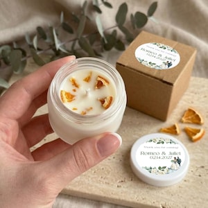 May include: A small glass jar candle with white wax topped with dried orange peel pieces. Beside it sits a brown cardboard box and a white lid, both featuring a circular label with floral graphics and the text Romeo & Juliet 02.14.2027.