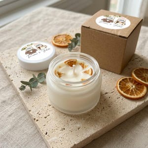 Puede incluir: Una vela blanca en un frasco de vidrio transparente decorada con rodajas de naranja seca y una mecha. A su lado hay una tapa blanca y una caja de cartón marrón, ambas con una pegatina circular que muestra el texto Romeo & Juliet y un dibujo.