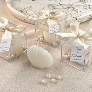 May include: Clear plastic boxes tied with ivory ribbons, each containing a white seashell-shaped candle and faux pearls. The boxes have small tags that read "Alicia & Mohamed 20 Juillet 2025". A loose candle and pearls are also present.